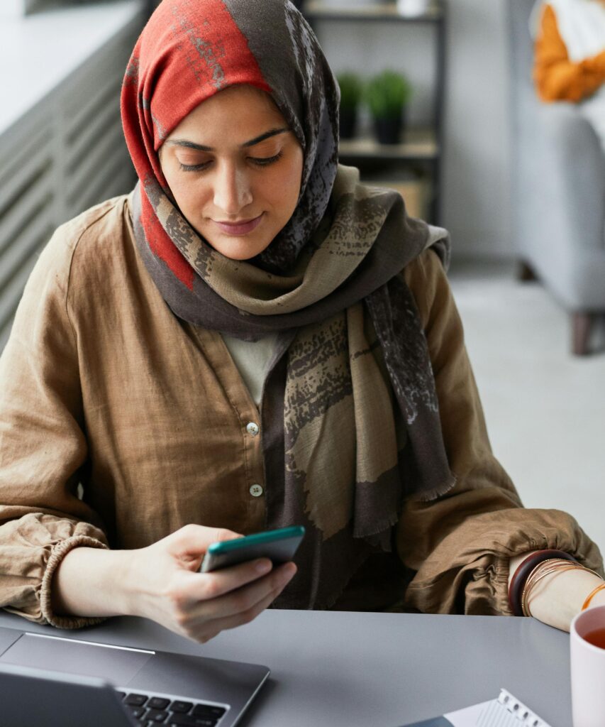 En kvinna sitter och tittar i sin mobil. Hon bär mönstrad hijab och brun blus.