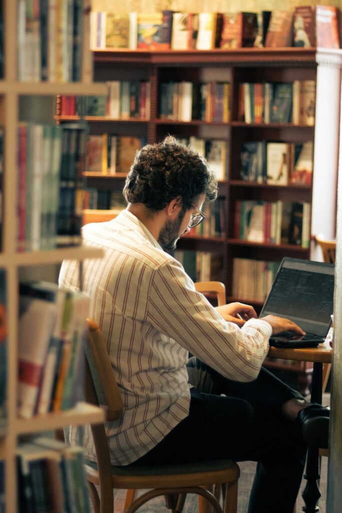 En man sitter på biblioteket och skriver på en dator. Han har mörkt hår, skägg och glasögon. Ryggen mot kameran.