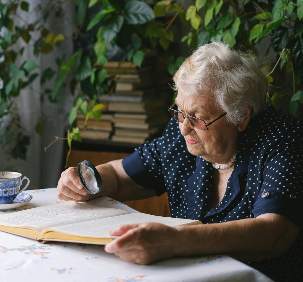 Äldre kvinna i glasögon och grått hår. Hon sitter vid bordet och läser med hjälp av ett förstoringsglas.