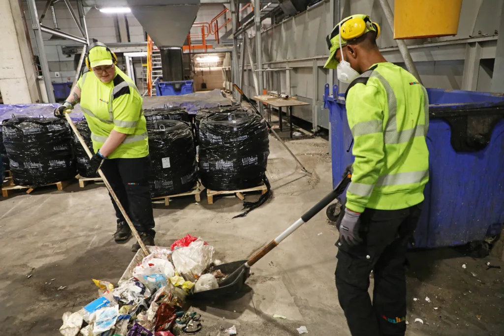 Två personer i gula arbetskläder. De sopar golvet inne på plaståtervinningen. 