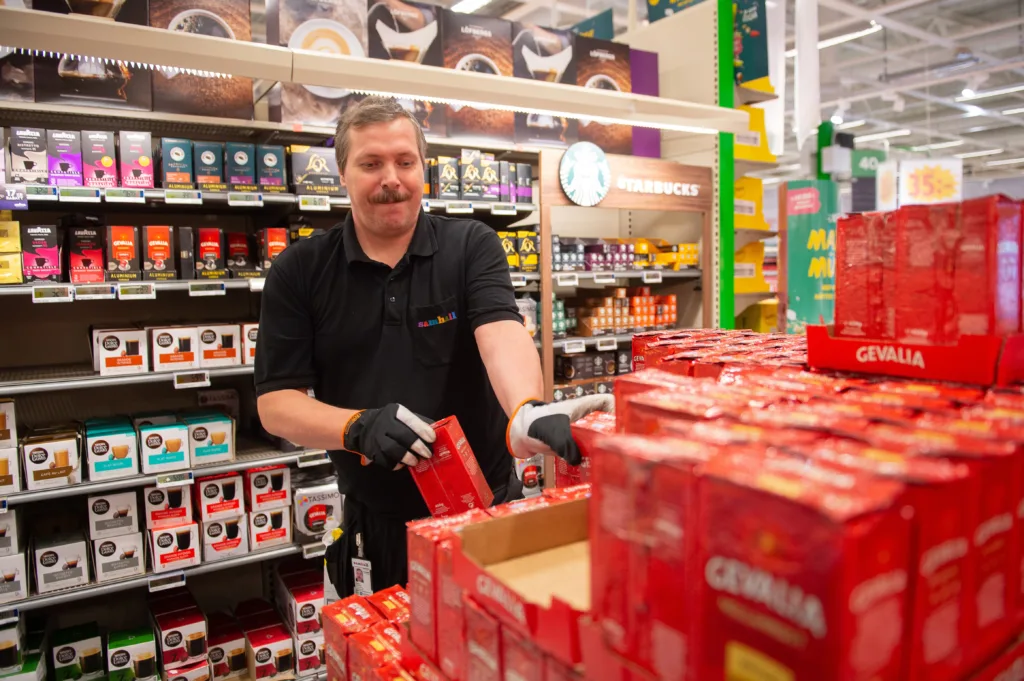 Man i svarta Samhallkläder och handskar. Han arbetar inne på Coop och står och packar upp kaffepaket.