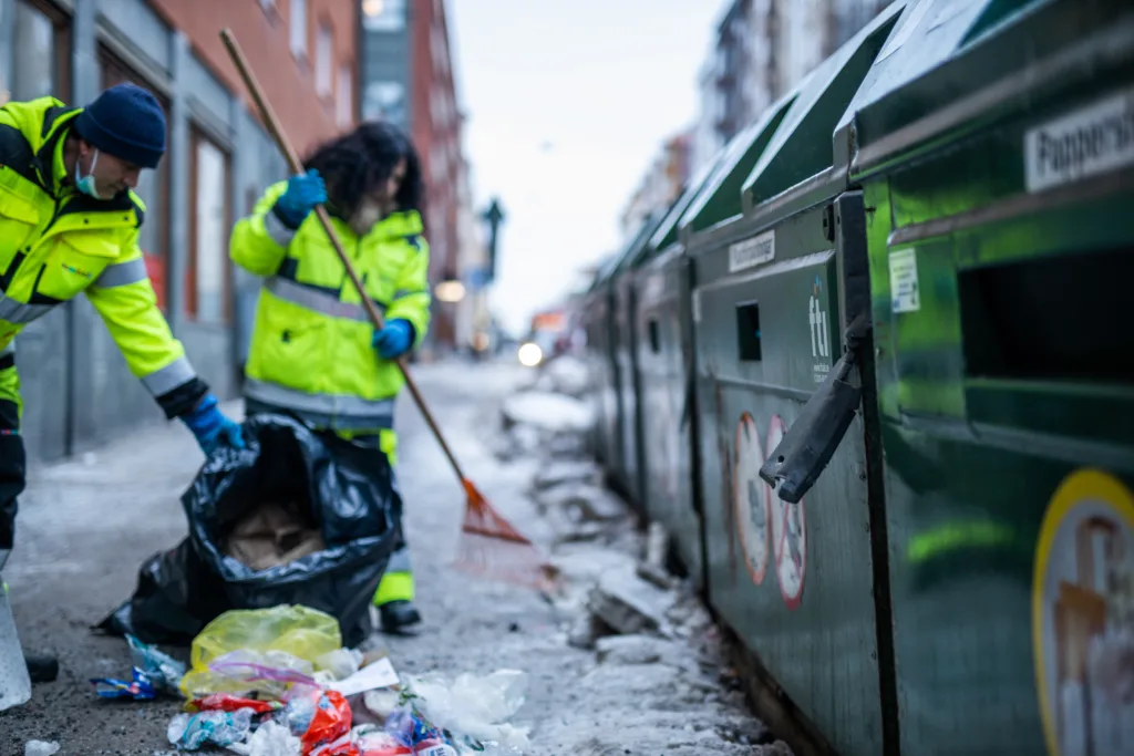 En man och en kvinna i gula Samhallkläder arbetar utomhus. De sopar vid en återvinningsstation och plockar upp avfall.