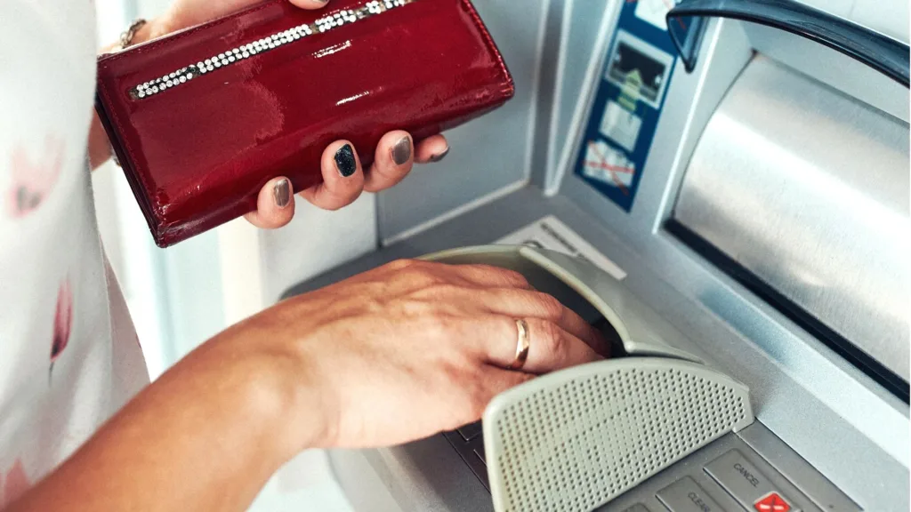 En kvinna tar ut pengar vid en bankomat. I vänsterhanden håller hon en röd plånbok. Med högerhanden knappar hon in uppgifter.