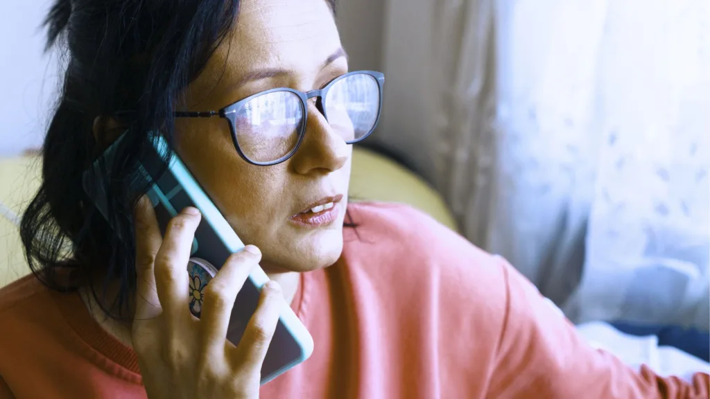 Kvinna i glasögon, mörkt hår och rosa tröja. Hon håller sin mobil mot örat.