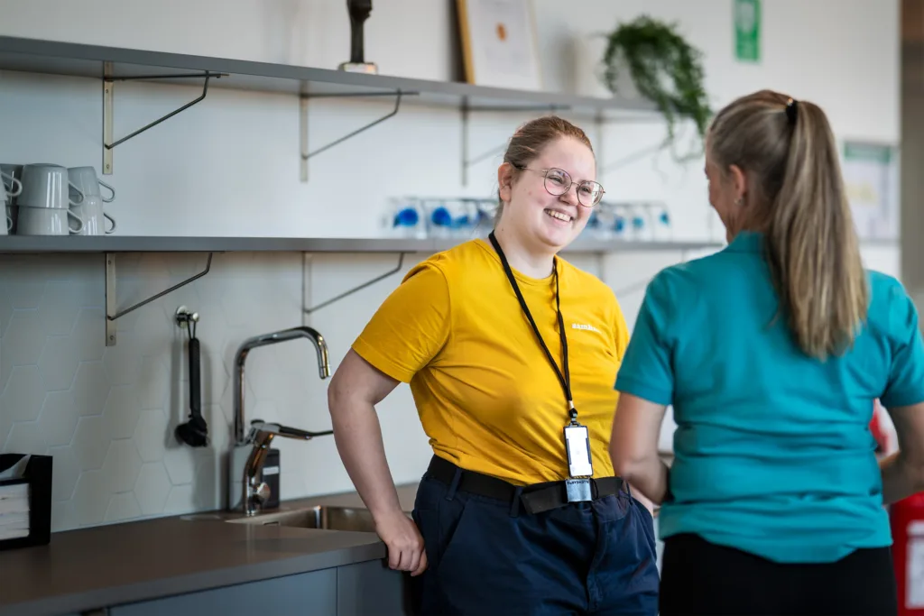 Tjej i gul Samhalltröja står och pratar med en kvinna som har ryggen mot kameran. De står vid en diskbänk. Tjejen i den gula tröjan skrattar.