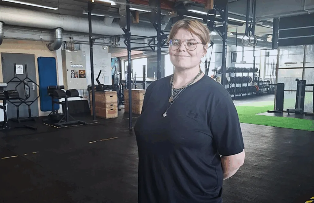 Kajsa Svensson på plats i gymmet. Hon har glasögon, flera silverhalsband och bär en mörk T-shirt.
