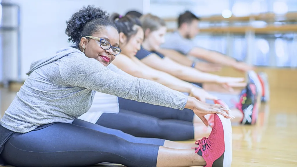 En grupp personer sitter på golvet i en gymlokal. De stretchar och sträcker sig mot sina fötter. Kvinnan närmast i bild ler och tittar in i kameran.