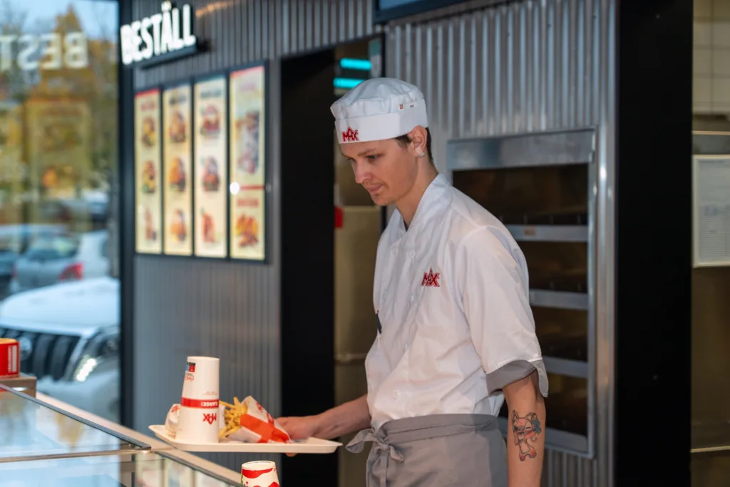 Kim Koskela i vita Max-kläder. Han står vid disken och håller i en bricka med hamburgare i papper.