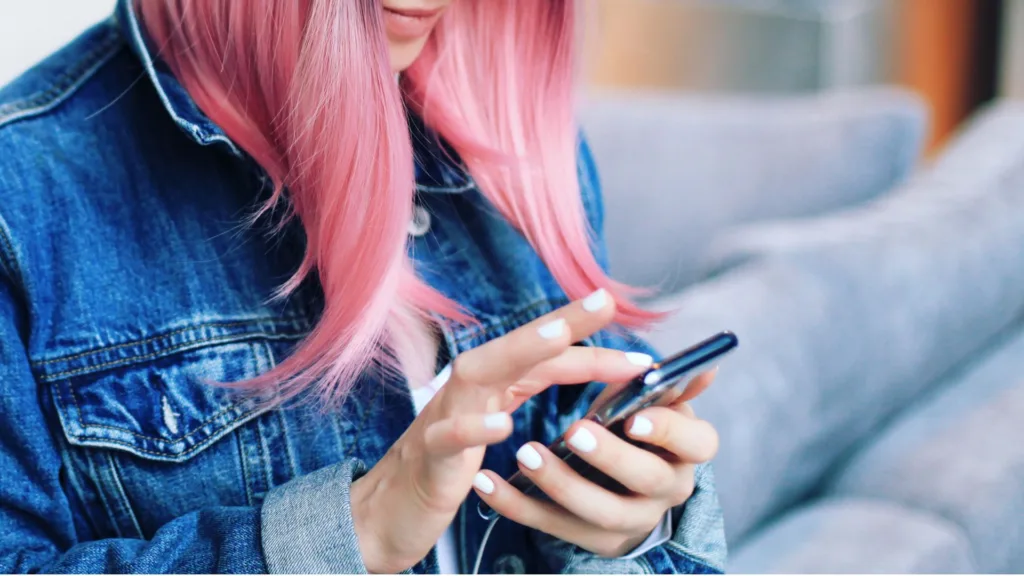 En person i rosa hår och jeansjacka står och läser i mobilen.